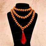 A double-strand beaded necklace made of rudraksha seed beads with a bright red tassel, displayed on a black jewelry bust against a beige stone-textured background.