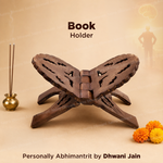 A dark brown wooden book holder shaped like the pages of an open book, displayed on a beige background with a blurred silhouette of a person and incense sticks in the background. Text overlays read 'Book Holder' and 'Personally Abhimanit by Dhwani Jain'.