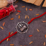 Handcrafted 925 silver Damru and Trishul Rakhi with red adjustable thread, inspired by Lord Shiva, displayed on a decorative background with kumkum, rice grains, and wooden accents. Traditional Raksha Bandhan bracelet, spiritual Hindu gift for brother, sacred Shiva rakhi for festival celebration.