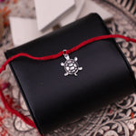 A rakhi bracelet with a small silver turtle charm, displayed across a black textured cylindrical holder on a patterned surface.