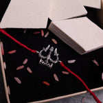 Handcrafted silver-tone Trishul and Damru Rakhi with an adjustable red thread, inspired by Lord Shiva, displayed on a neutral gray background with decorative rice grains and white stone props. Traditional Hindu Raksha Bandhan bracelet, spiritual brother gift, Shiva Trident Rakhi for festive celebration and devotional wear.