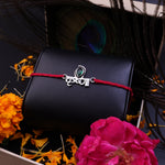 A red cord bracelet with a silver-tone 'krishna' charm and a green stone accent, displayed on a black textured box surrounded by orange flowers, a pink rose, and peacock feathers.