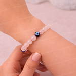 Pink beaded bracelet with a blue evil eye charm on a wrist against a light pink background.