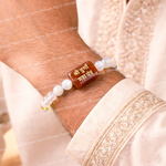 Bracelet with white beads and a brown rectangular stone on a wrist, worn by a person in traditional attire.