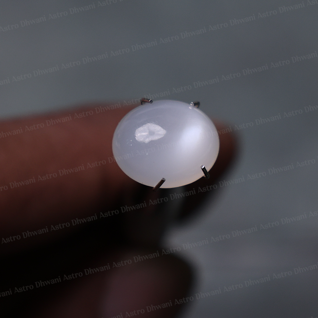 A close-up of a small oval white cabochon gemstone held in a metal prong setting