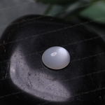 A small round, dome-shaped white gemstone with a smooth, polished surface, resting on a dark, textured stone. The background is dark gray/black with a soft-focus green leaf in the upper right corner.