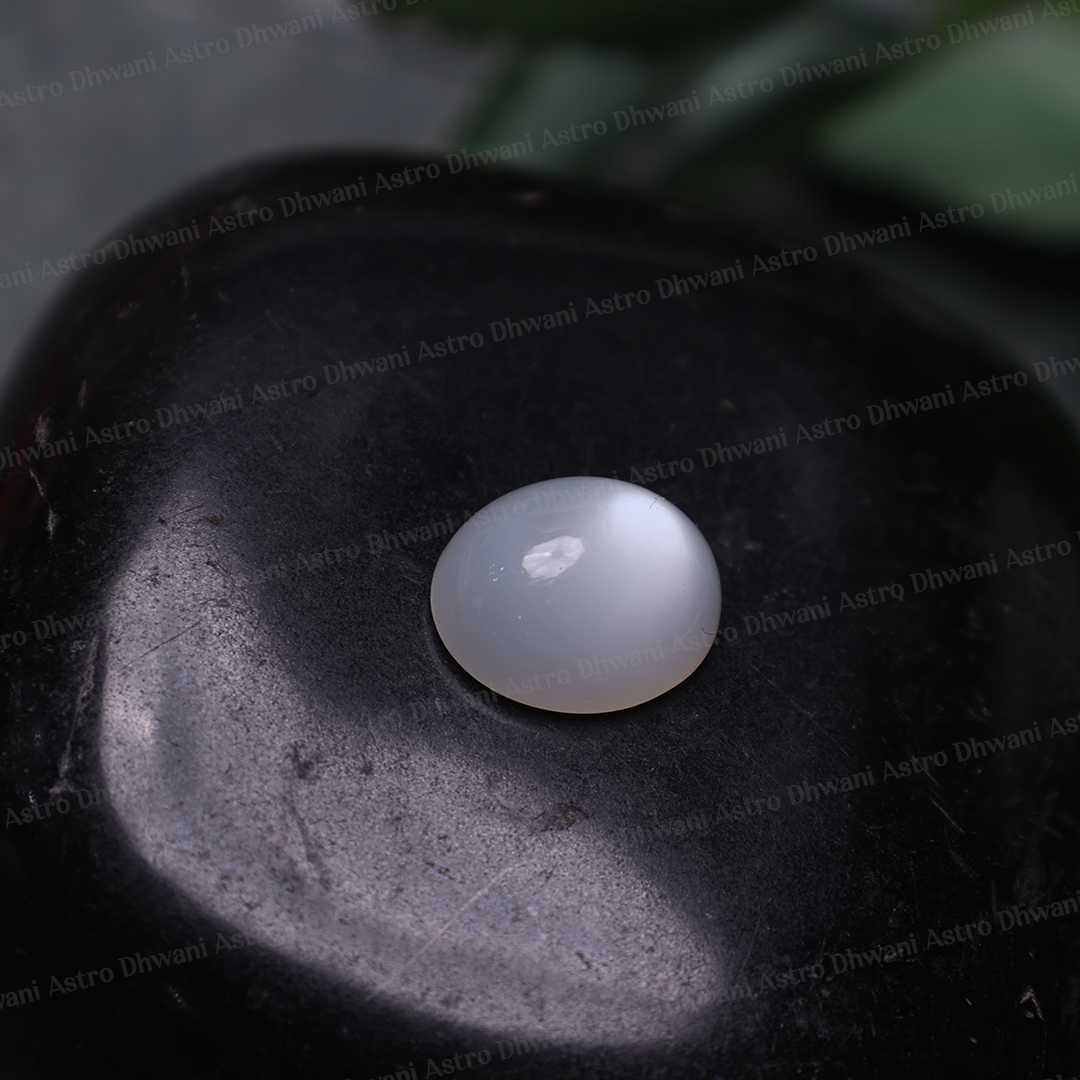 A small round, dome-shaped white gemstone with a smooth, polished surface, resting on a dark, textured stone. The background is dark gray/black with a soft-focus green leaf in the upper right corner.Opal Gemstone – Spiritual Healing Gemstone | Loose Opal Stone for Jewelry, Meditation & Crystal Collection