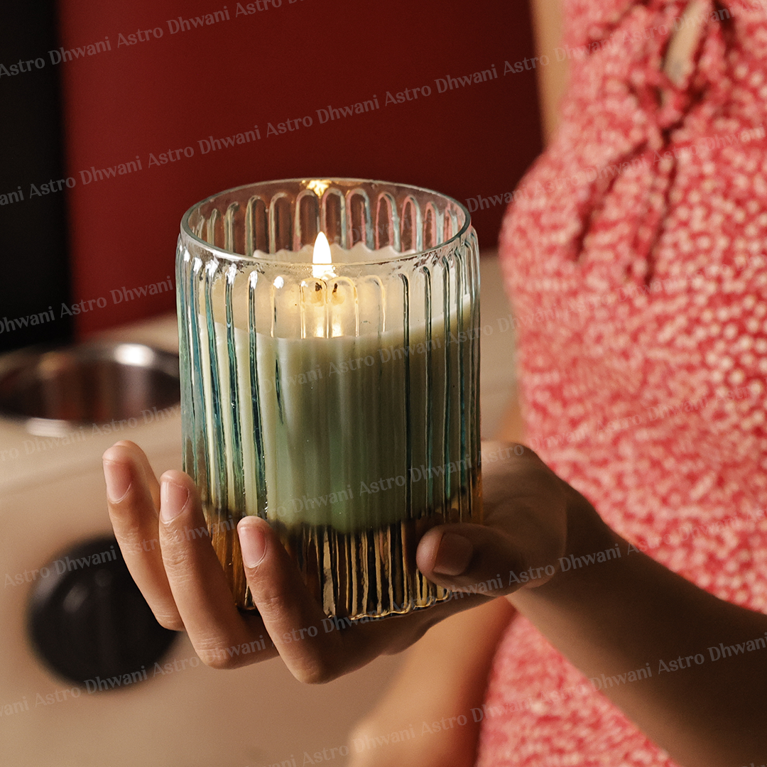 A hand holds a lit scented candle in a ribbed clear glass holder with a warm gold-toned base, set against a softly blurred indoor background with a person wearing a red patterned top.