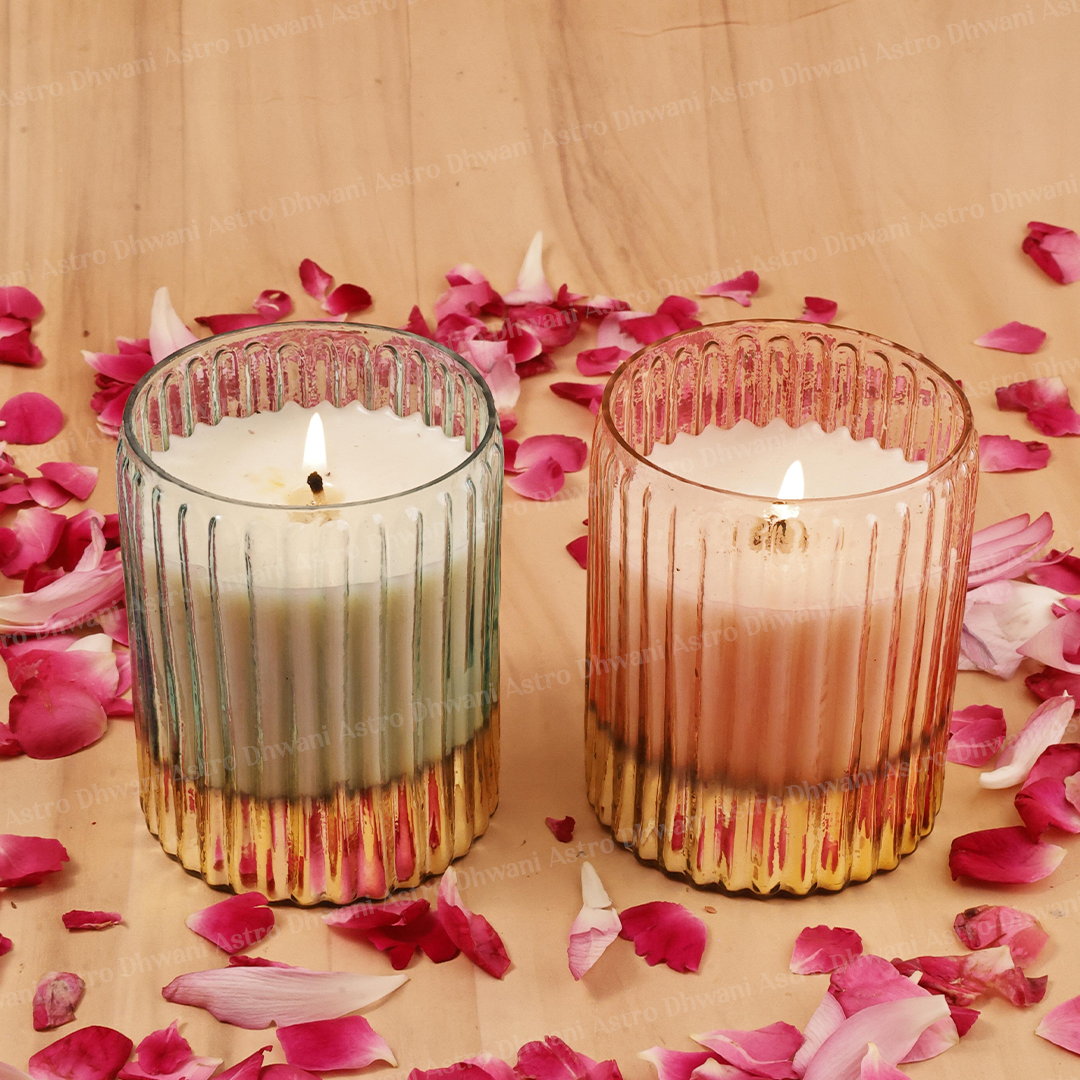 Two lit candle holders in ribbed glass with pink and gold accents, placed on a wooden surface surrounded by scattered pink flower petals.