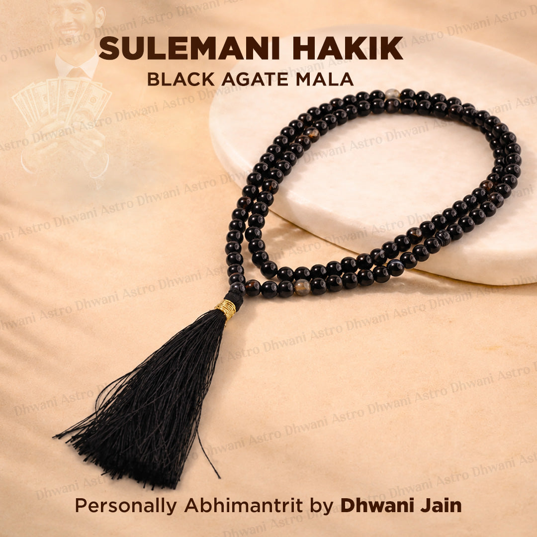 Black agate mala necklace with a black tassel and a small gold accent bead, displayed on a beige fabric background with a white stone slab. Text overlays read 'SULEMANI HAKIK BLACK AGATE MALA'.