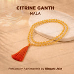 A citrine gemstone mala necklace with a vibrant orange tassel, displayed on a white stone slab over a beige fabric background.