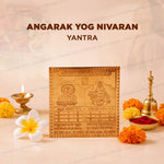 A square metallic gold-toned yantra plaque with embossed Hindu deity illustrations and Sanskrit text, displayed on a beige surface with a diya, brass bell, red powder, marigold flowers, and a white flower. Large text at the top reads 'ANGARAK ROG NIVARAN YANTRA'.
