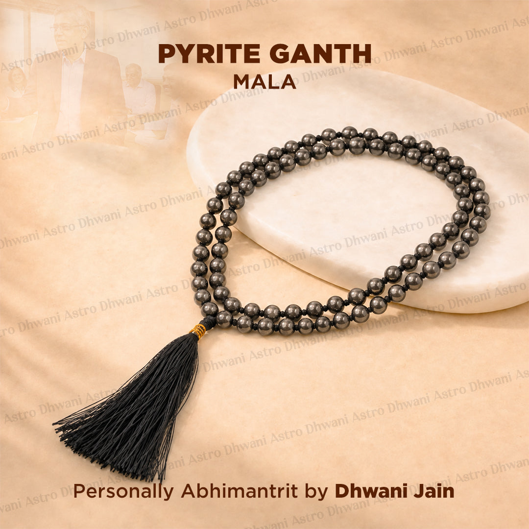 A dark gray pyrite bead mala necklace with a black tassel, displayed on a white stone slab over a beige fabric background with text overlays.