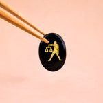 Black stone with gold Libra zodiac symbol held by tweezers on a peach background