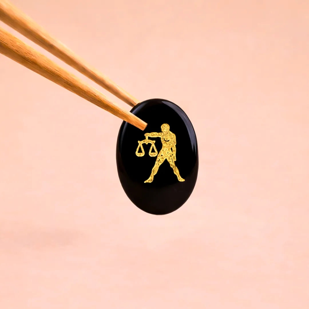 Black stone with gold Libra zodiac symbol held by tweezers on a peach backgroundLibra Zodiac Sign Coin – Abhimantrit Astrology Coin for Balance & Relationships