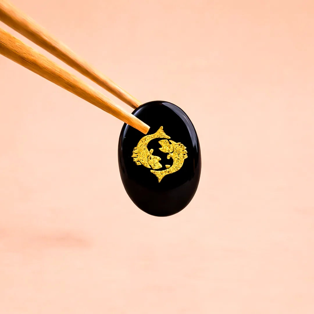 Black stone with gold symbol held by chopsticks on a peach backgroundPisces Zodiac Sign Coin – Abhimantrit Astrology Coin for Peace