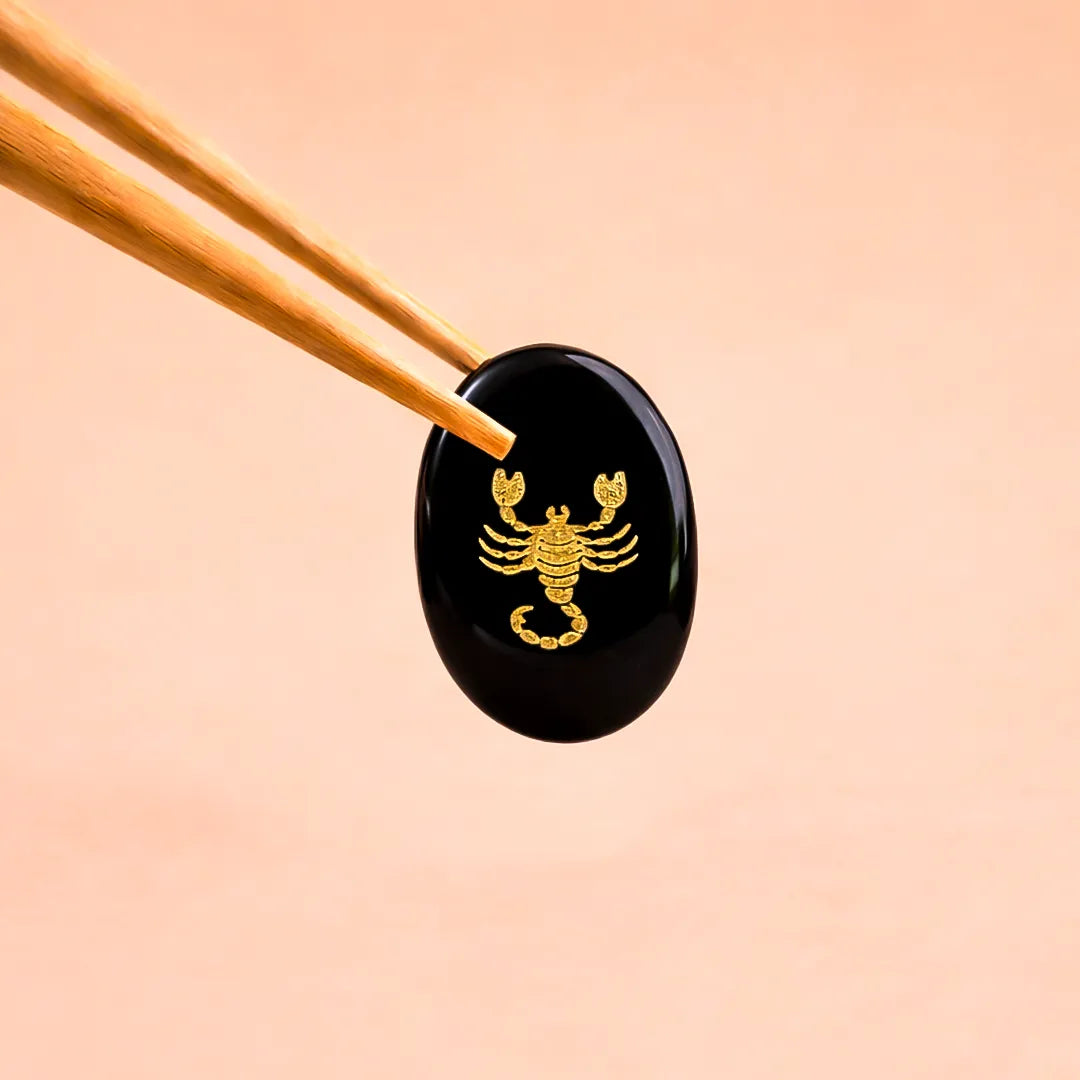 Black stone with gold scorpion design held by chopsticks on a peach background