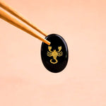 Black stone with gold scorpion design held by chopsticks on a peach background