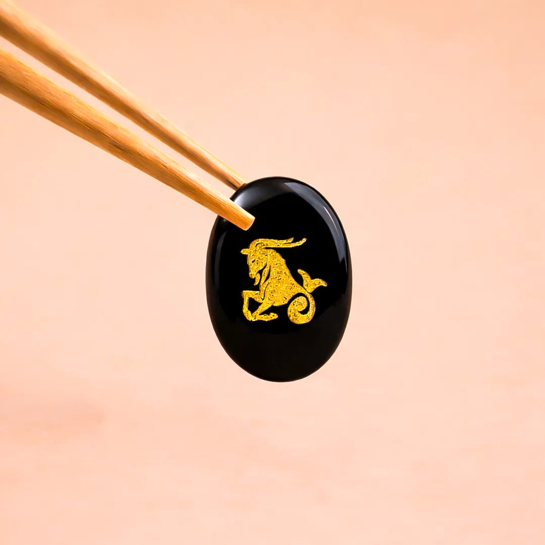 Black stone with gold deer engraving held by chopsticks on a peach backgroundCapricorn Zodiac Sign Coin – Abhimantrit Astrology Coin for Stability