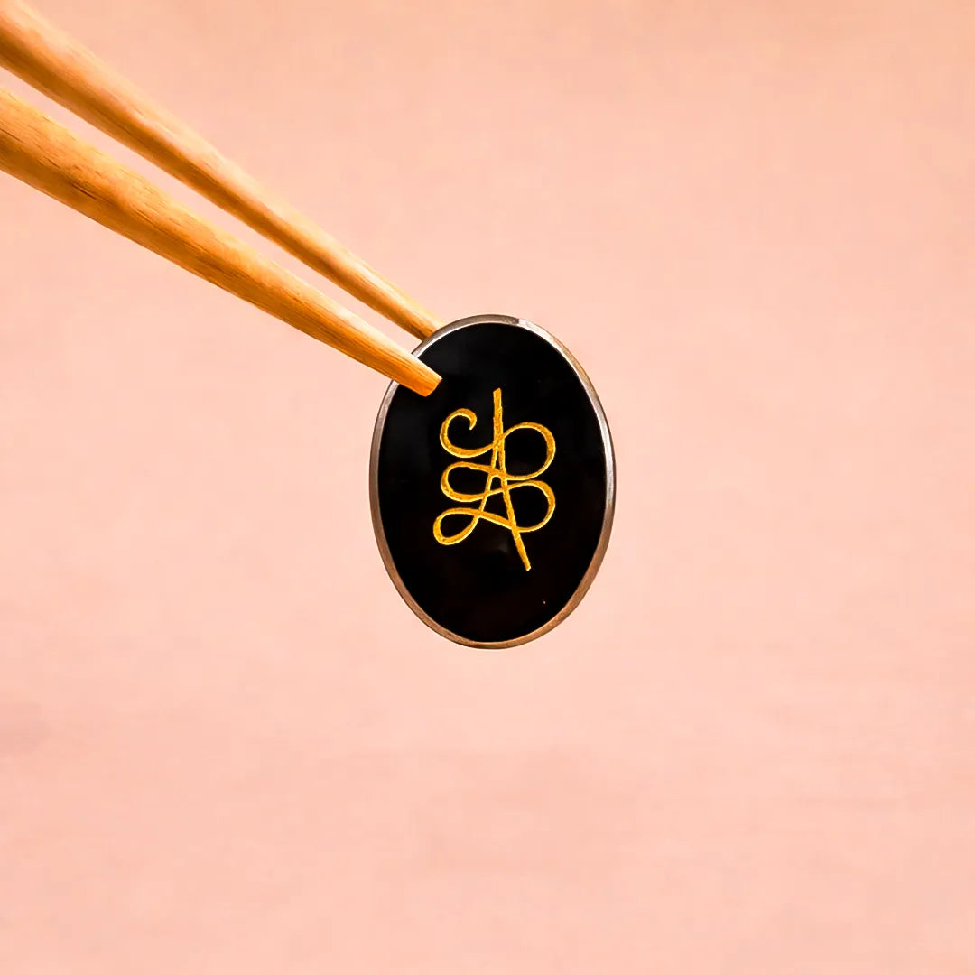 Black and gold circular object with a logo on a pink backgroundPyrite Zibu Coin – Abhimantrit for Wealth Protection