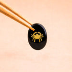 Black stone with gold crab design held by chopsticks on a beige background