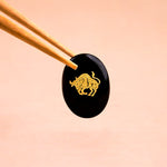 Black stone with gold bull engraving held by chopsticks on a peach background