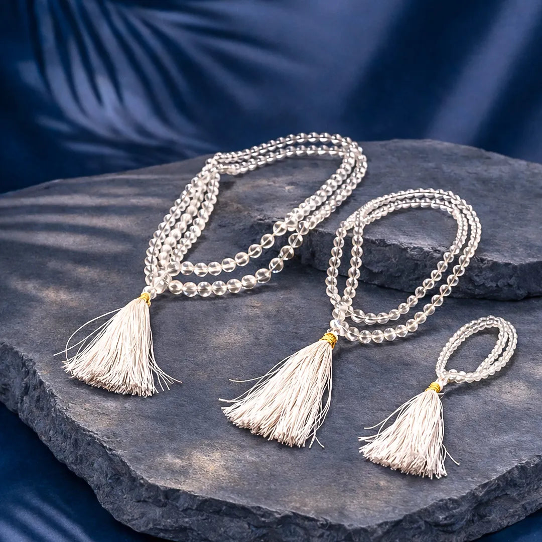 Three crystal necklaces with tassels on a dark stone surface against a blue fabric backgroundClear Quartz Crystal Mala – Master Healer Meditation