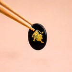 Black stone with gold lion design held by chopsticks on a beige background