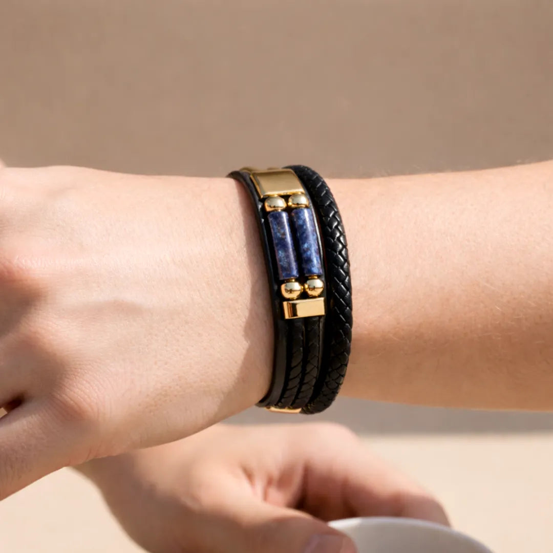 Black braided leather bracelet with blue stones and gold accents on a wrist against a beige background