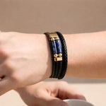 Black braided leather bracelet with blue stones and gold accents on a wrist against a beige background