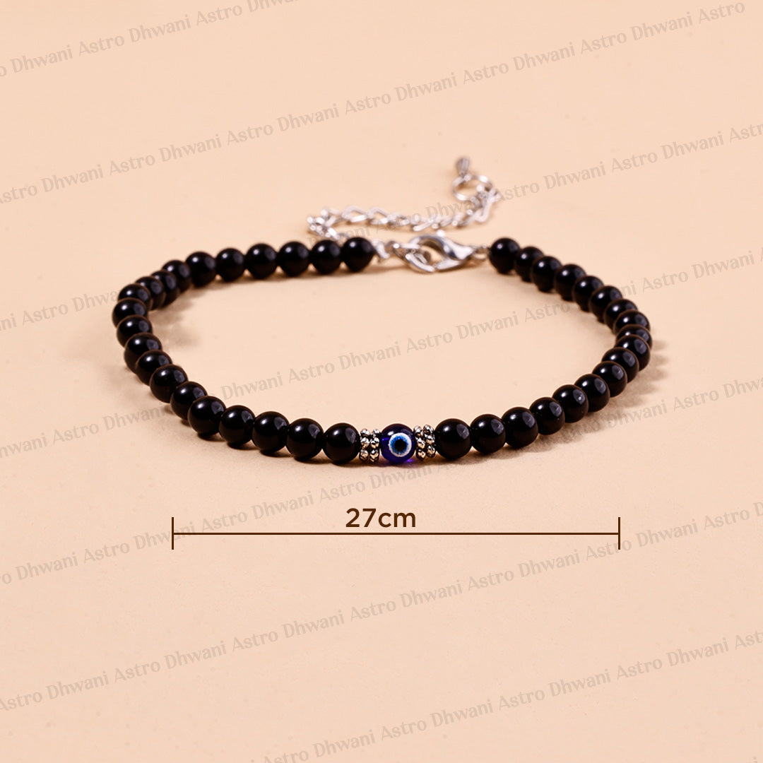A black beaded bracelet with a central evil eye charm and silver-tone clasp, shown on a beige background with a 27cm size indicator.