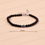 A black beaded bracelet with a central evil eye charm and silver-tone clasp, shown on a beige background with a 27cm size indicator.