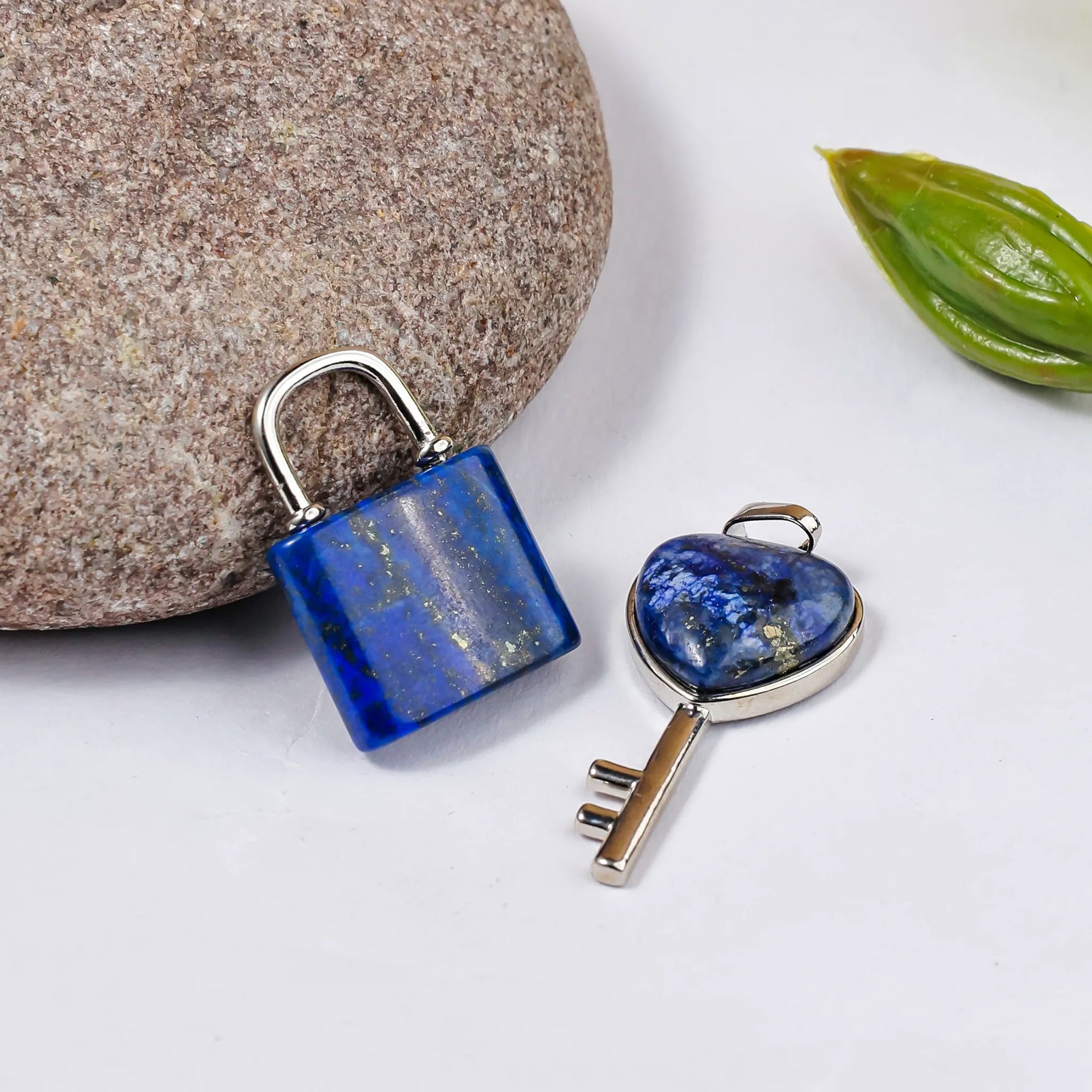 Blue padlock and key on a white surface with a stone and leaf in the backgroundLapis Lazuli Lock And Key Pendant - Abhimantrit & Certified