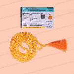 A set of natural citrine (yellow) glass prayer beads with an orange tassel, displayed next to a printed certificate card on a solid peach-pink background.