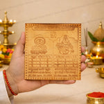 A hand holds a square wooden Hindu calendar plaque featuring Lord Shiva and Goddess Lakshmi, with Devanagari text and a swastika symbol at the top, set against a warm, decorated background with brass lamps and red powder.