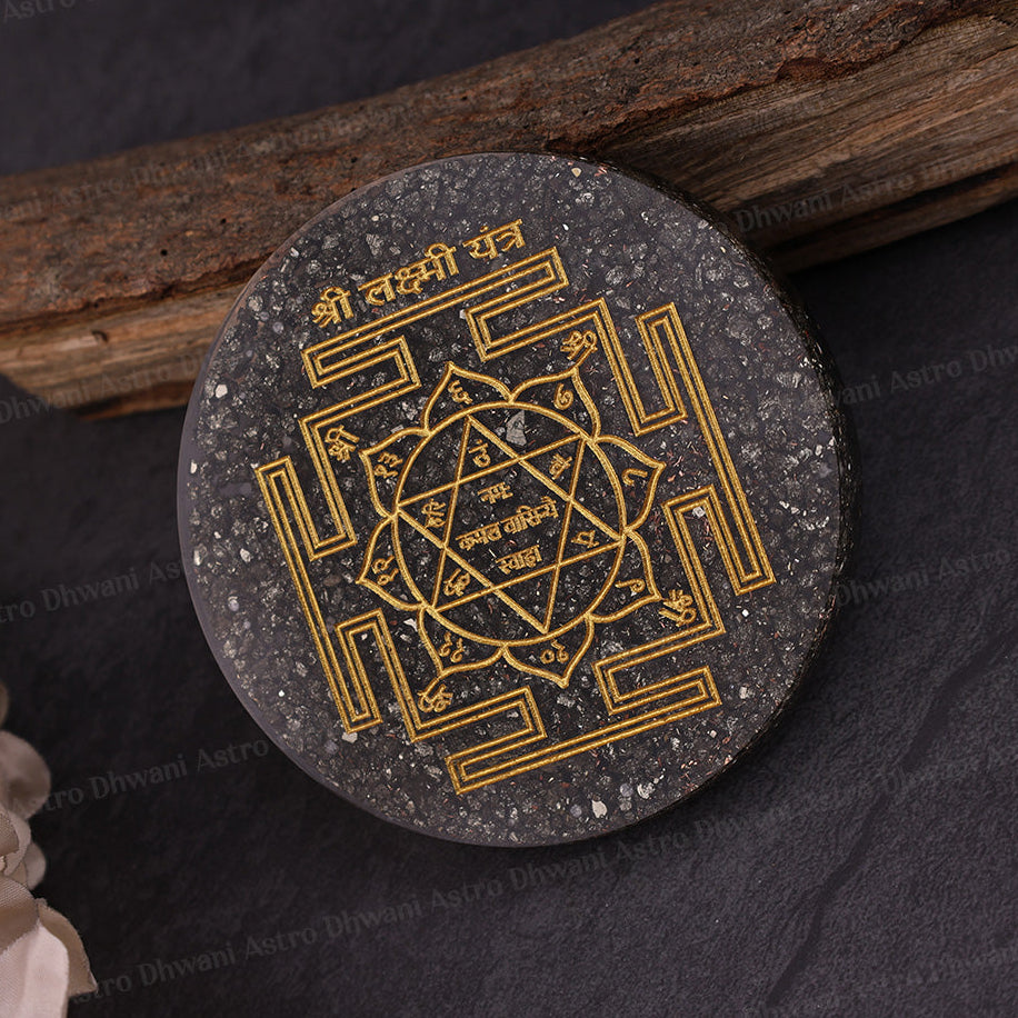 A round black and gray stone yantra plaque with gold-colored engraved geometric symbols and Sanskrit text, displayed on a dark textured surface with a piece of wood and pale flowers as props.