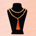 A double-strand beaded necklace with a large orange tassel pendant, displayed on a black velvet jewelry bust against a light peach-pink background 