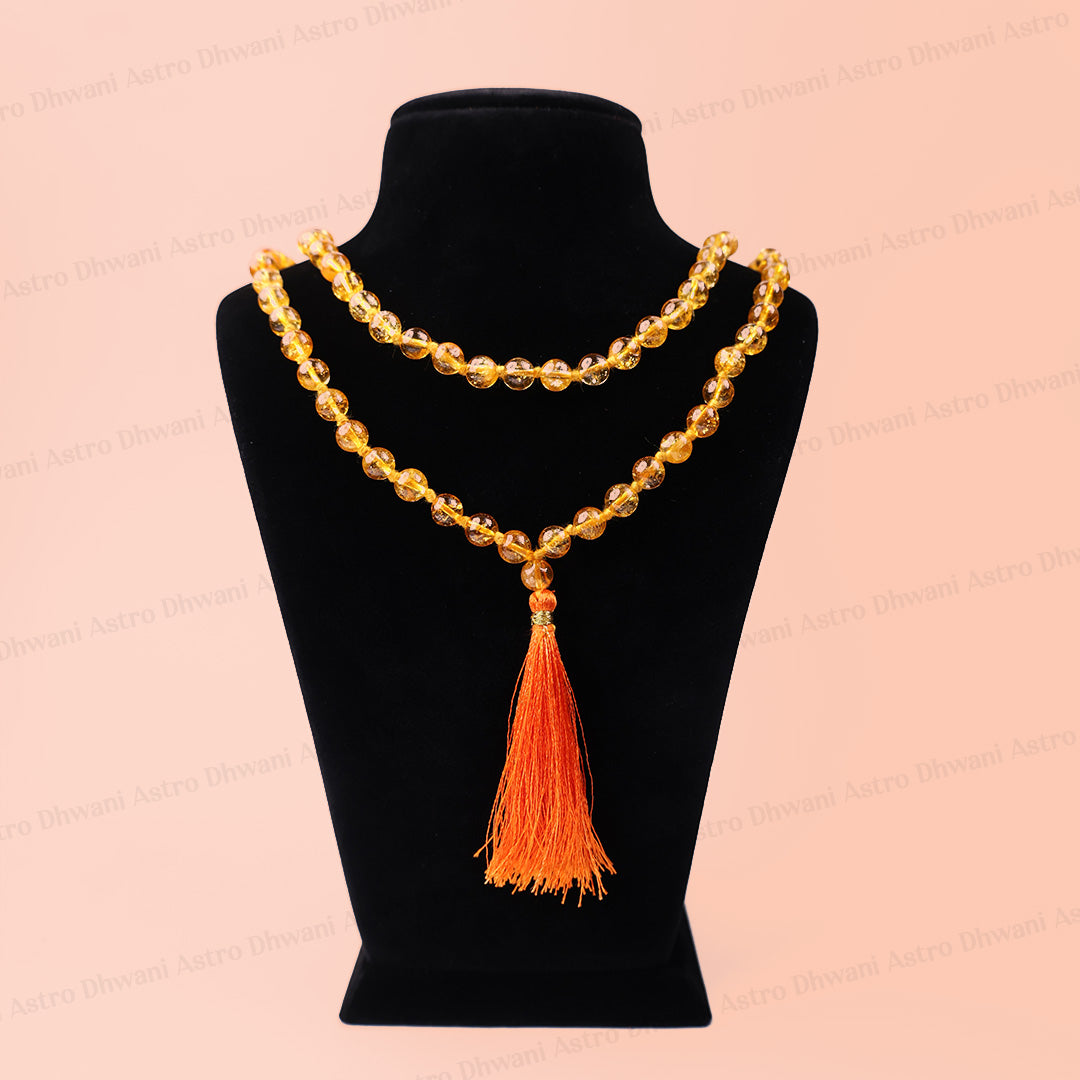 A double-strand beaded necklace with a large orange tassel pendant, displayed on a black velvet jewelry bust against a light peach-pink background Natural Yellow Citrine Ganth Mala | Prosperity & Abundance Mala | Handknotted Japa Mala for Meditation, Puja & Spiritual Practice