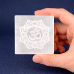 Hand holding a square crystal with Om symbol design against a dark blue background