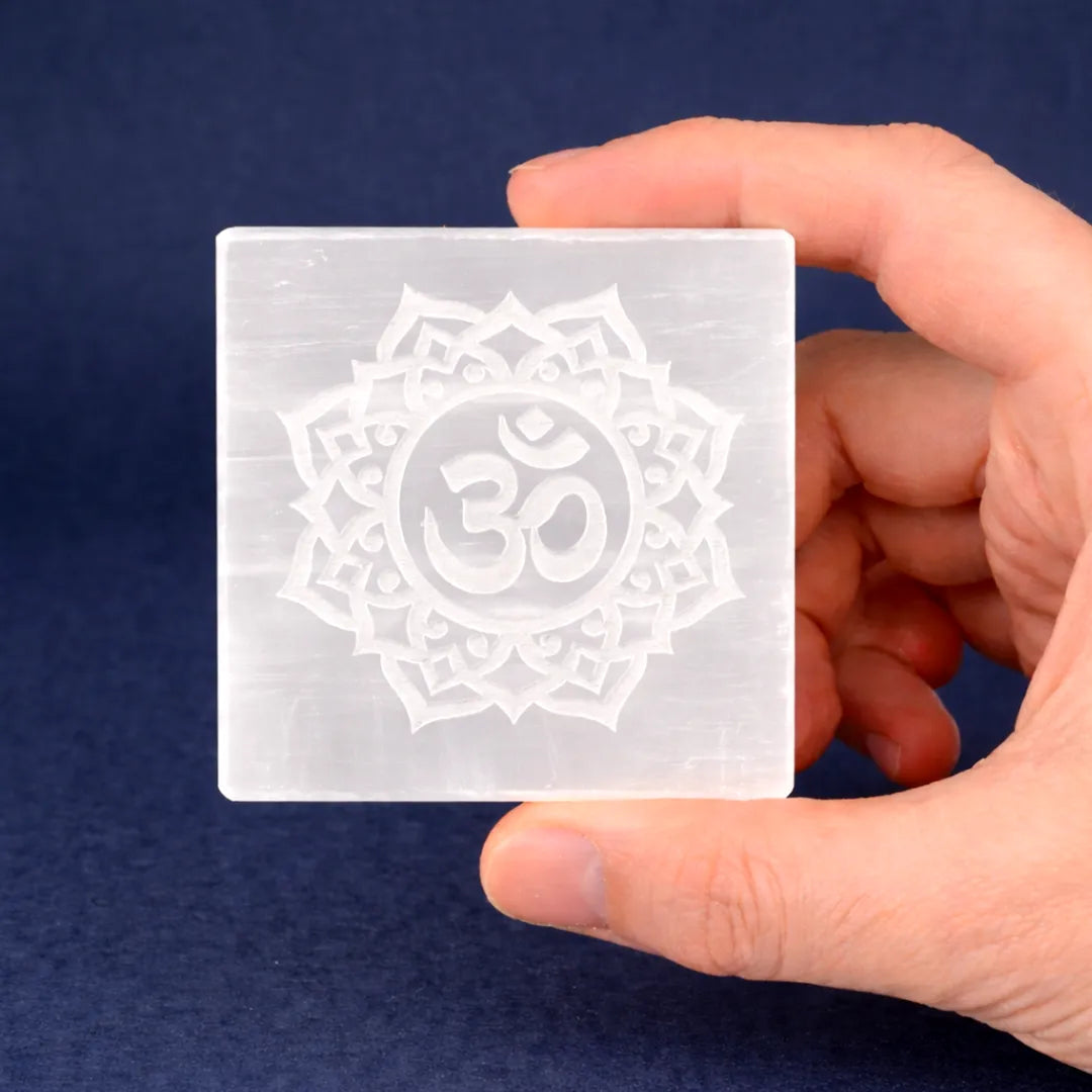 Hand holding a square crystal with Om symbol design against a dark blue backgroundOM Selenite Plate – Square Energy Cleansing & Charging Plate