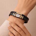 Black braided leather bracelet with gold and silver accents on a wrist against a beige background