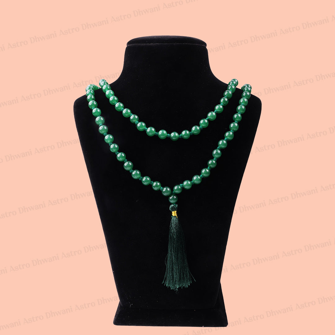 A green aventurine beaded necklace with a tassel pendant displayed on a black velvet jewelry bust against a peach-pink background.