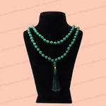 A green aventurine beaded necklace with a tassel pendant displayed on a black velvet jewelry bust against a peach-pink background.