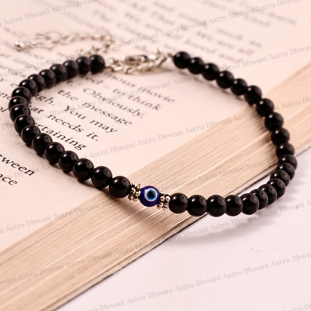 A beaded black stone bracelet with a blue and white evil eye charm and silver-toned metal accents, displayed on an open book.Black Obsidian Anklet | Evil Eye Protection Anklet | Positivity & Negative Energy Shield