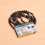A coiled strand of dark natural sapphire beads with a matching paper certificate card, displayed on a light peach background.