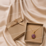 Gold necklace with a purple pendant in a brown box on a beige fabric background