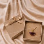 Gold necklace with a purple pendant in an open box on a beige fabric background