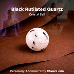 Black rutilated quartz crystal ball on a reflective surface with blurred people in the background