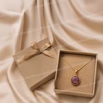 Gold necklace with a purple pendant in a brown box on a beige fabric background