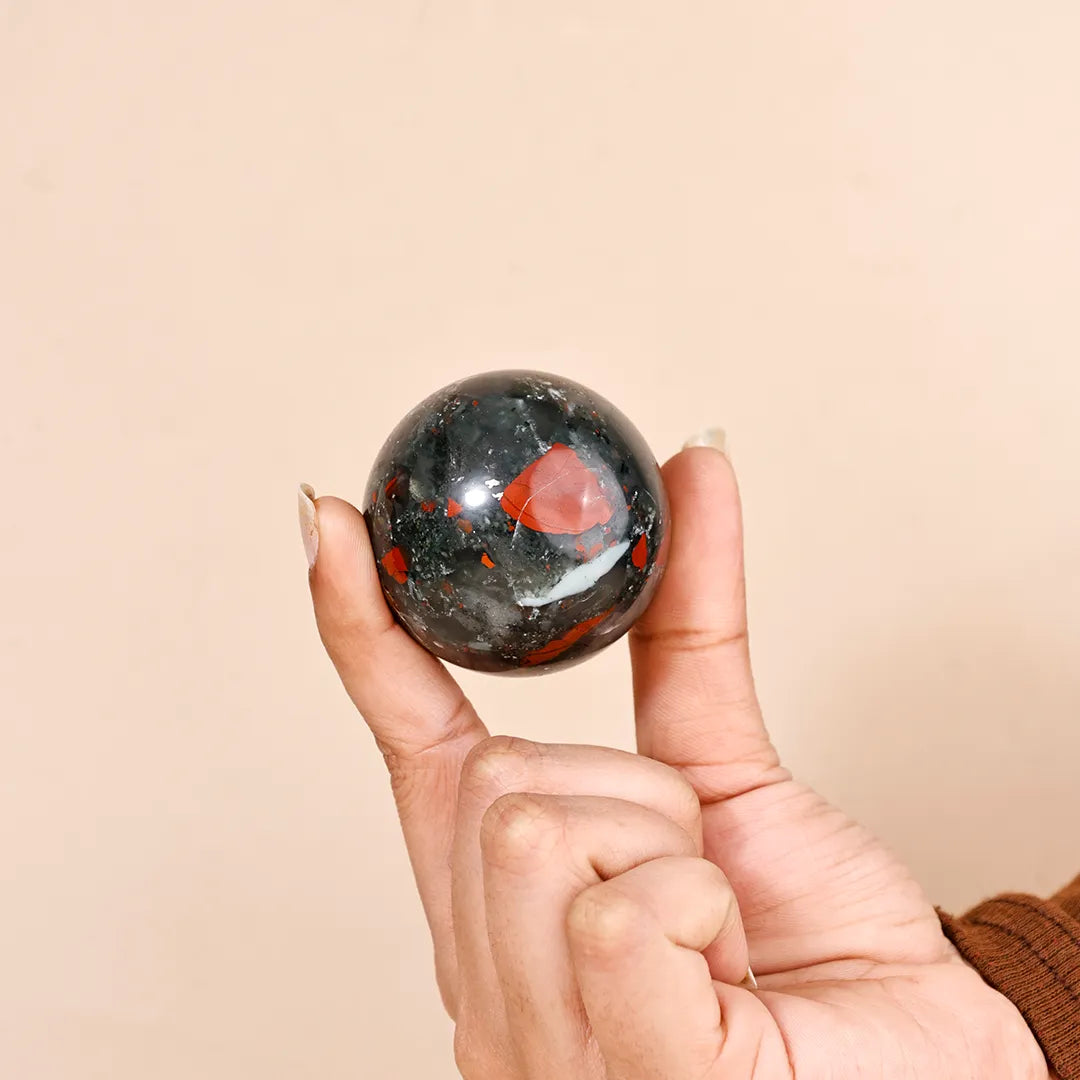 Hand holding a bloodstone crystal ball against a beige backgroundNatural Blood Stone Crystal Ball – Abhimantrit & Certified